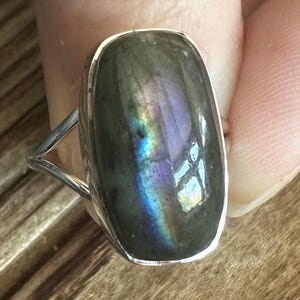 May include: A silver ring featuring a large, oval-shaped labradorite gemstone. The gemstone has a dark grey base with iridescent blue, green, and purple flashes.