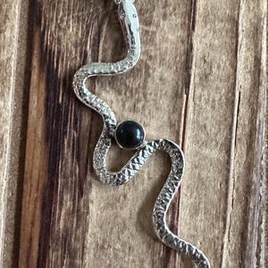 May include: A silver snake pendant with a black gemstone. The snake is curled in a S shape and has a textured surface. The pendant has a loop at the top for attaching to a chain.