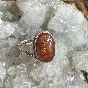 May include: A silver ring with a large oval-shaped sunstone gemstone. The stone is a reddish-brown color with flecks of gold.