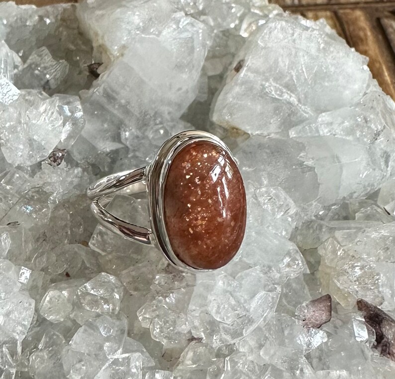 May include: A silver ring with a large oval-shaped sunstone gemstone. The stone is a reddish-brown color with flecks of gold.