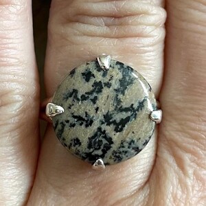 May include: A silver ring with a round, black and brown speckled stone set in the center. The stone is surrounded by four silver prongs.