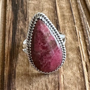 May include: A silver ring featuring a pink and white gemstone set in a teardrop shape. The ring has a braided design around the edge of the setting.