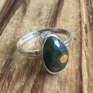 May include: A silver ring featuring an oval-shaped, faceted gemstone. The gemstone is primarily dark green with a central yellow and brown pattern. The ring band has a textured design.