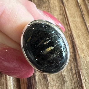 May include: Oval-shaped ring featuring a large, polished black stone with gold and white inclusions, set in a silver-colored metal band. The ring's design is simple, highlighting the natural beauty of the stone.