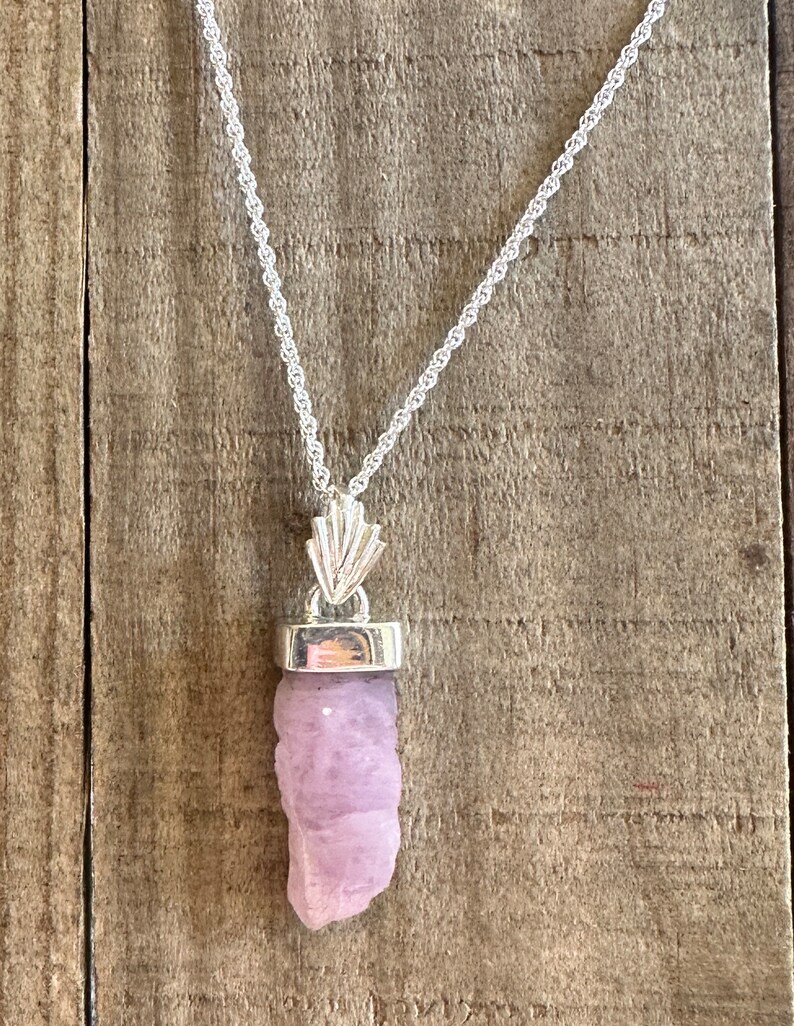 Puede incluir: Un collar de cadena de plata con un colgante que presenta un cristal crudo de color p&uacute;rpura claro. El cristal est&aacute; engastado en un bisel de plata y cuelga de una argolla de plata.