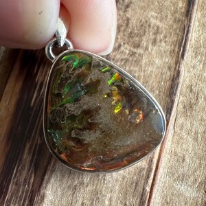 May include: A silver pendant featuring a teardrop-shaped iridescent opal with green and brown hues. The opal has a unique, swirling pattern.