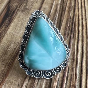 May include: A large, triangular, polished gemstone ring. The stone is a light blue-green color with white marbling. The ring has a decorative, ornate silver setting with a scrolled design around the stone.
