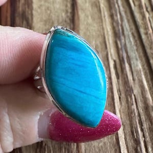 May include: A silver ring featuring a large, marquise-shaped turquoise stone. The stone has a vibrant blue color with visible natural patterns. The ring's setting is a polished silver, enhancing the stone's color. The ring is a statement piece.