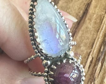 May include: A silver ring with a teardrop-shaped moonstone and a round, pink gemstone. The ring is set with small silver beads around the stones.