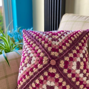 May include: A crocheted cushion with a geometric pattern in shades of pink, white, and brown. The cushion has a button in the centre.