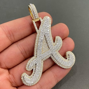 May include: A gold pendant in the shape of the letter A, encrusted with small, clear diamonds. The pendant is on a chain and is being held by a hand.