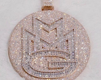 May include: A gold pendant with a round, diamond-encrusted design featuring the letters "MG" in a stylized font.