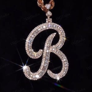 以下が含まれることがあります： きらびやかなクリアクリスタルを多数ちりばめた、大きな「B」の文字が特徴のローズゴールドのペンダントネックレス。ペンダントは、お揃いのロープチェーンから吊り下げられています。背景は黒です。