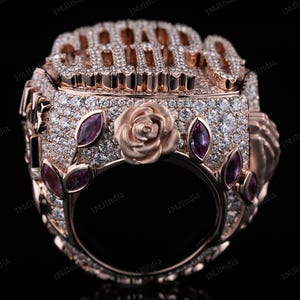 May include: A rose gold ring with a large, diamond-encrusted inscription that reads "CHAMP" and a rose design on the band. The ring is adorned with small pink gemstones.