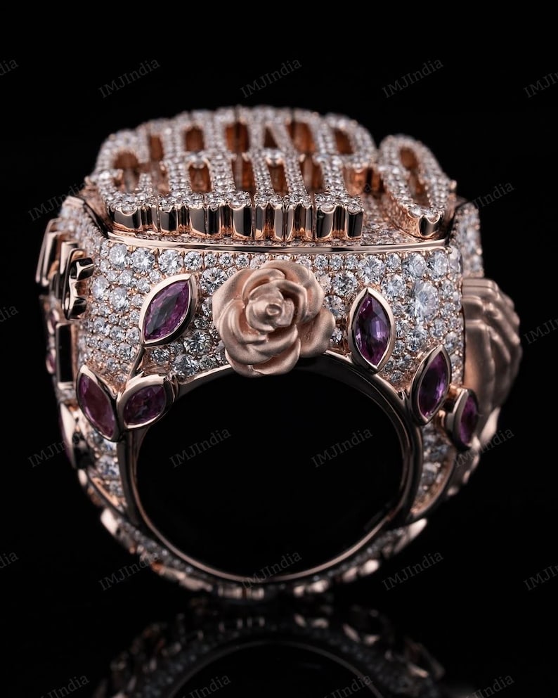 May include: A rose gold ring with a large, diamond-encrusted inscription that reads "CHAMP" and a rose design on the band. The ring is adorned with small pink gemstones.