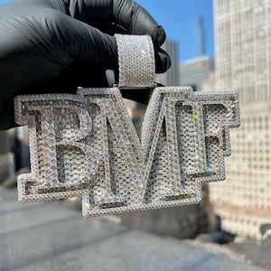 Può includere: Un ciondolo d'argento con le lettere "BMF" incastonate con numerose piccole pietre scintillanti. Il ciondolo è tenuto contro uno sfondo di un paesaggio urbano sotto un cielo azzurro. Anche l'anello del ciondolo è ricoperto di pietre.