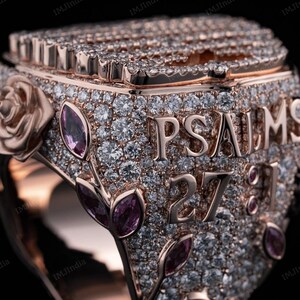 May include: A rose gold ring with a square face, covered in small diamonds and pink gemstones. The ring says "PSALMS 27:1" in raised letters.