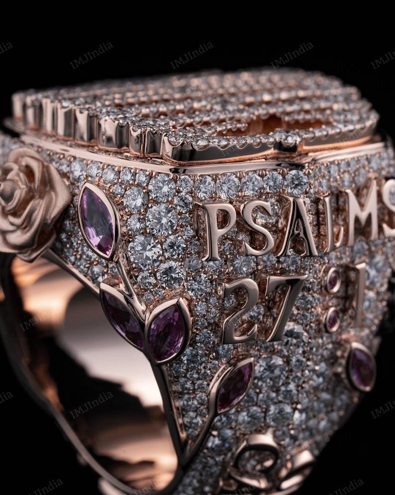 May include: A rose gold ring with a square face, covered in small diamonds and pink gemstones. The ring says "PSALMS 27:1" in raised letters.