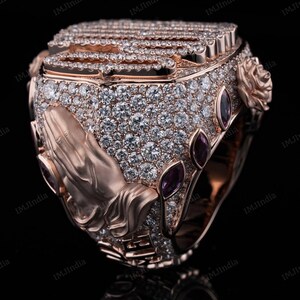 May include: A rose gold ring with a praying hands design, set with clear and purple gemstones. The ring is intricately detailed and features a textured surface.