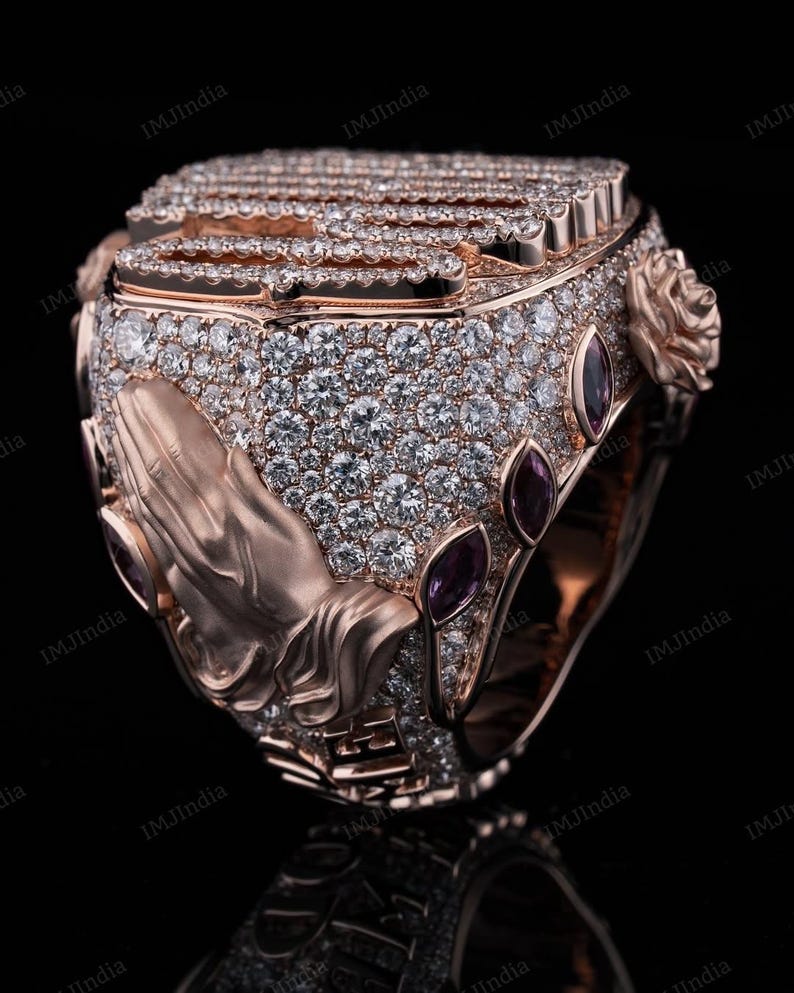 May include: A rose gold ring with a praying hands design, set with clear and purple gemstones. The ring is intricately detailed and features a textured surface.