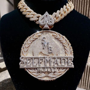May include: A gold chain necklace with a large pendant featuring a diamond-encrusted circle with the words "SELF MADE" and a spade symbol. The pendant is surrounded by a wreath of leaves.