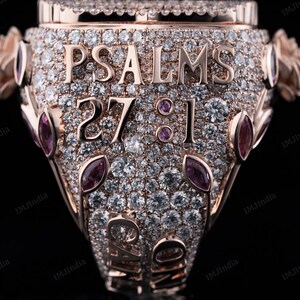 May include: A rose gold ring with a large, clear stone in the center. The ring is covered in smaller, clear stones and has the words "PSALMS 27:1" engraved on the band.