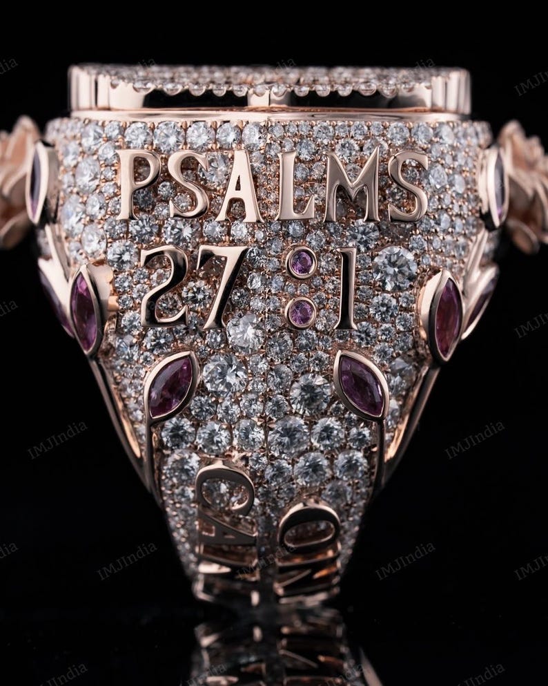 May include: A rose gold ring with a large, clear stone in the center. The ring is covered in smaller, clear stones and has the words "PSALMS 27:1" engraved on the band.