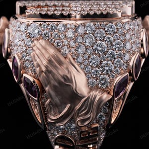 May include: A gold ring with a praying hands design, set with small diamonds and accented with purple gemstones.