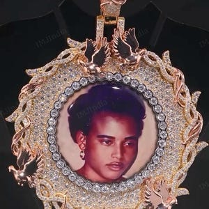May include: A gold-coloured pendant with a portrait set within a diamond-encrusted frame. The frame is decorated with ornamental details and small bird figures. The pendant has a diamond-covered bail and the text "IMJIndia" is visible.