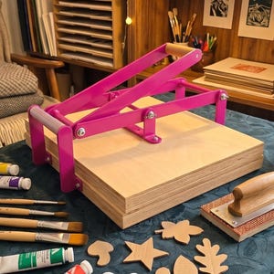 May include: A bright pink manual bookbinding press with wooden boards, surrounded by several paint tubes, wooden-handled paintbrushes, a wooden brayer, and various small cardboard cutouts in shapes like stars and leaves on a dark green tablecloth.