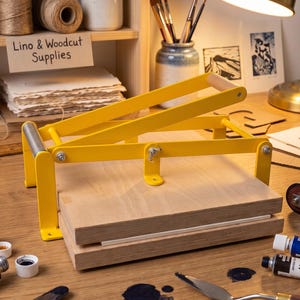 May include: A yellow metal lino and woodcut printing press featuring a lever mechanism and two stacked wooden boards. The press is positioned on a workspace surrounded by paint tubes, a brayer, and art supplies.