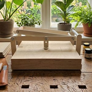 May include: A light-colored wooden bookbinding press with a lever mechanism, positioned on a rustic wooden workbench. Beside the press are several metal carving tools in a leather roll, a stack of paper, and small glass jars of colored ink.