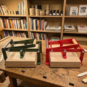 May include: Two bookbinding presses, one in dark green and one in bright red, each featuring a metal frame with a wooden handle and a thick wooden base plate. The mechanical presses are designed with adjustable wing nuts and metal rods.