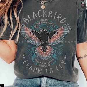 May include: Dark grey t-shirt featuring a black bird graphic with red and blue details, and the words "BLACKBIRD" and "LEARN TO FLY". The tee has a vintage, worn appearance.