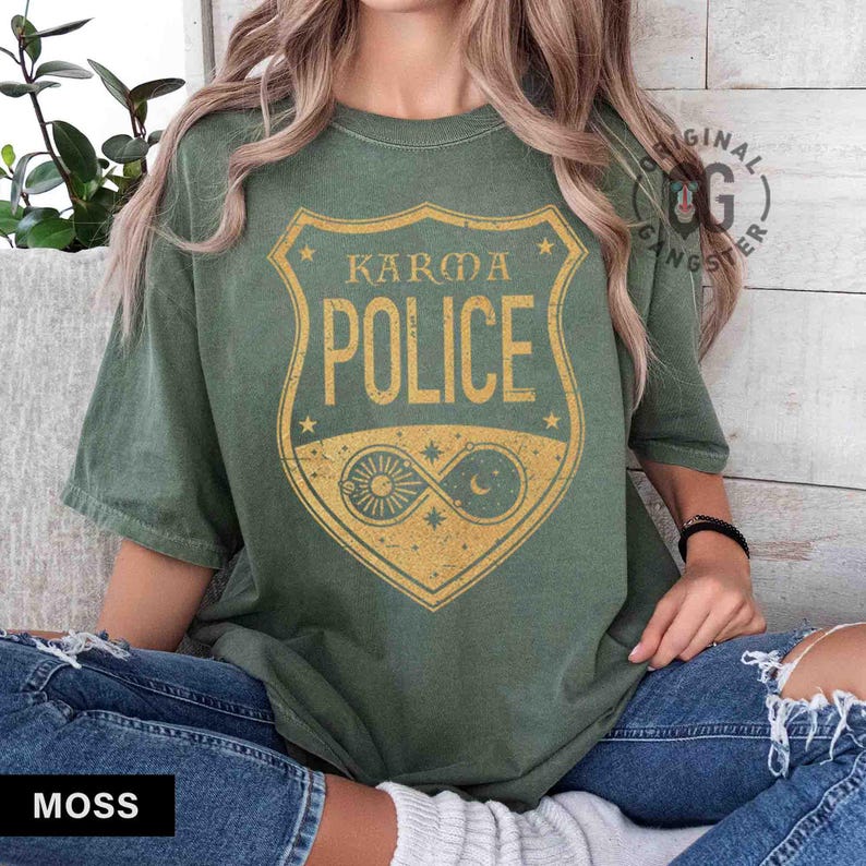May include: Moss green t-shirt with a distressed gold graphic of a shield that reads "KARMA POLICE". The shield design includes a sun, moon, and stars. The shirt is a relaxed fit.