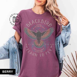 May include: Berry-colored t-shirt with a graphic of a blackbird with outstretched wings. The text on the shirt reads "BLACKBIRD" and "LEARN TO FLY". The shirt has rolled-up sleeves and a relaxed fit.