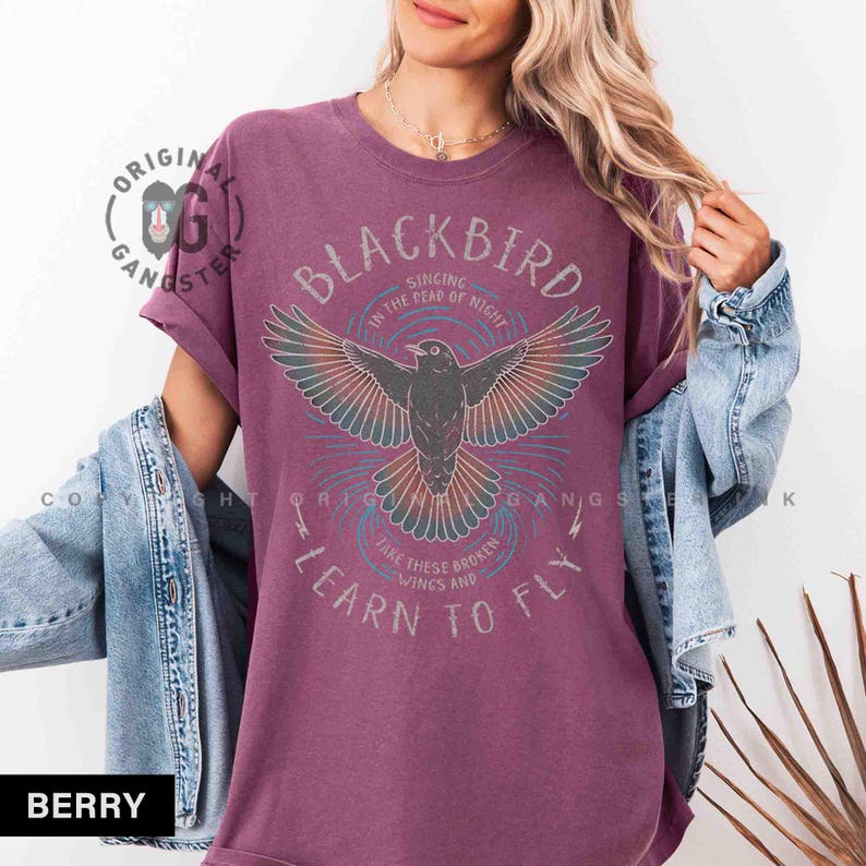 May include: Berry-colored t-shirt with a graphic of a blackbird with outstretched wings. The text on the shirt reads "BLACKBIRD" and "LEARN TO FLY". The shirt has rolled-up sleeves and a relaxed fit.
