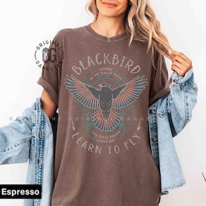 May include: Espresso-colored t-shirt featuring a graphic of a blackbird with outstretched wings, surrounded by text that reads "BLACKBIRD" and "LEARN TO FLY." The design includes red, teal, and white accents, creating a vintage aesthetic.