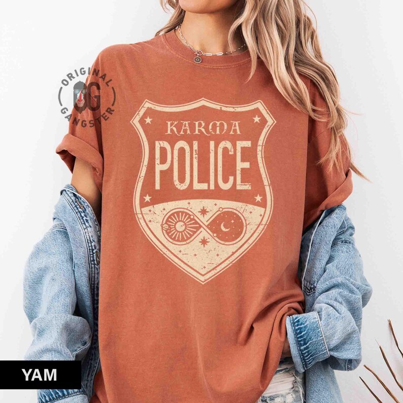 May include: Burnt orange t-shirt with a cream-colored graphic that reads "Karma Police" inside a shield design. The graphic includes a sun and moon symbol. The shirt has rolled-up sleeves and a relaxed fit.