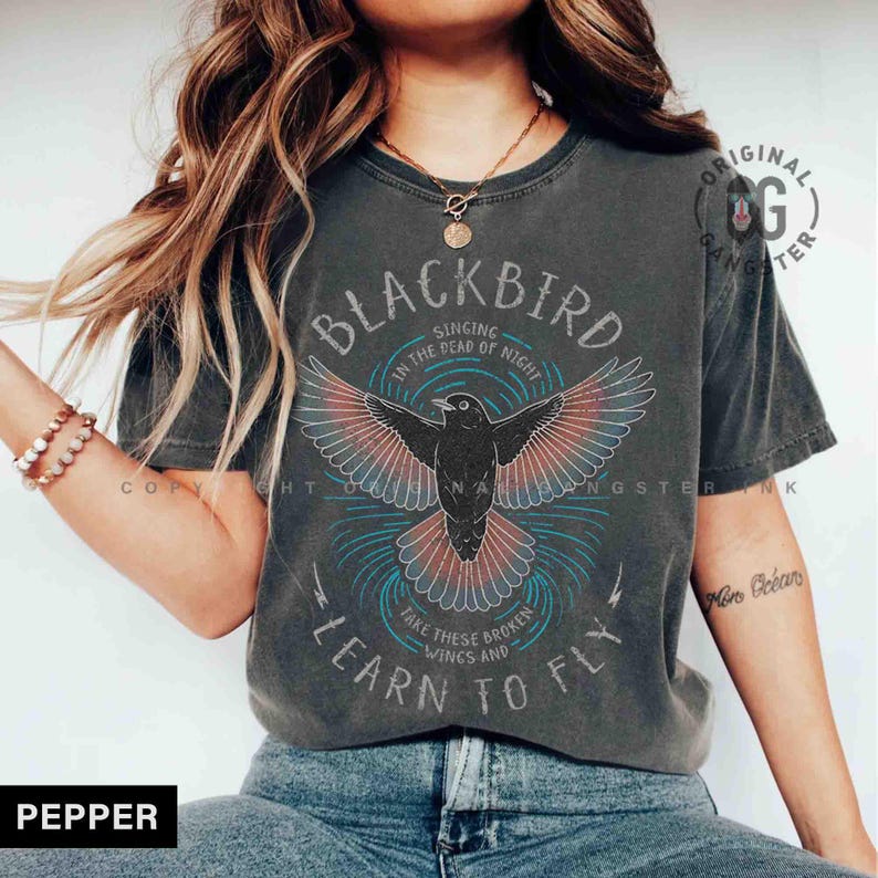 May include: Dark grey t-shirt featuring a black bird graphic with red and blue details, and the words "BLACKBIRD" and "LEARN TO FLY". The tee has a vintage, worn appearance.