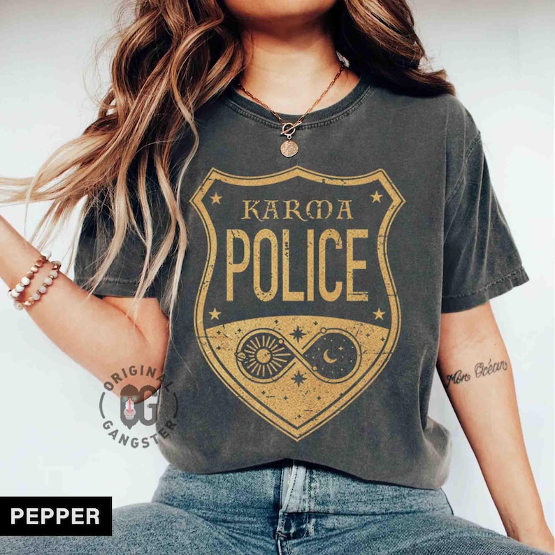 May include: Dark gray t-shirt with a distressed gold graphic that reads "KARMA POLICE" inside a shield design. The graphic includes celestial elements like the sun, moon, and stars. The shirt is a casual style.