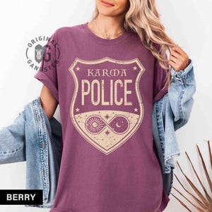 May include: Berry-colored t-shirt featuring a shield design with the words "KARMA POLICE" in a vintage style. The shield includes celestial elements like the sun, moon, and stars. The shirt has short sleeves and a relaxed fit.