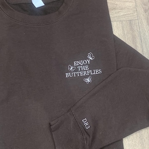 May include: A brown sweatshirt featuring white embroidered text and butterfly motifs that read "ENJOY THE BUTTERFLIES". The sleeve also has "DR3" embroidered in white.