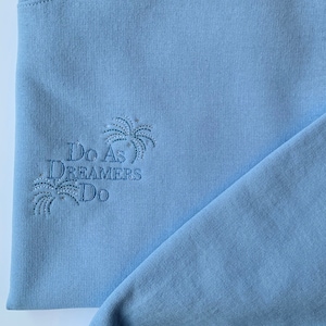 Op de afbeelding: Een lichtblauw sweatshirt met de tekst "Do As Dreamers Do" geborduurd op de voorkant. De tekst wordt omringd door een gestileerd palmboomontwerp.