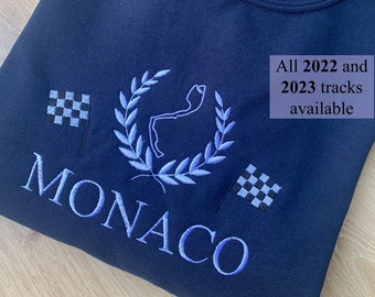 以下が含まれることがあります： 青い刺繍でレースコースを囲む月桂樹の輪、青い刺繍で「MONACO」の文字、そして2つの市松模様の旗が施された黒いスウェットシャツ。 「2022年と2023年のすべてのトラックが利用可能」というテキストも青い刺繍で施されています。