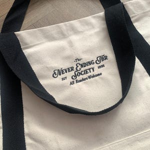 May include: Cream-colored tote bag with black handles and the text "-The- Never Ending TBR Society EST 1995 All Readers Welcome" printed in black. The bag has a simple, classic design.
