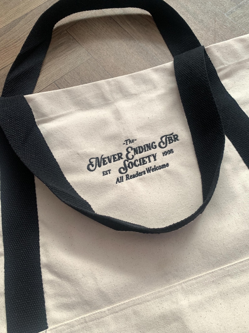 May include: Cream-colored tote bag with black handles and the text "-The- Never Ending TBR Society EST 1995 All Readers Welcome" printed in black. The bag has a simple, classic design.