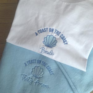 Puede incluir: Camiseta blanca y sudadera azul claro con dise&ntilde;os de conchas bordadas y las frases "A Toast on the Coast" y "Bride" en la camiseta y "That's Amore" en la sudadera. El texto y la concha son de color azul claro.