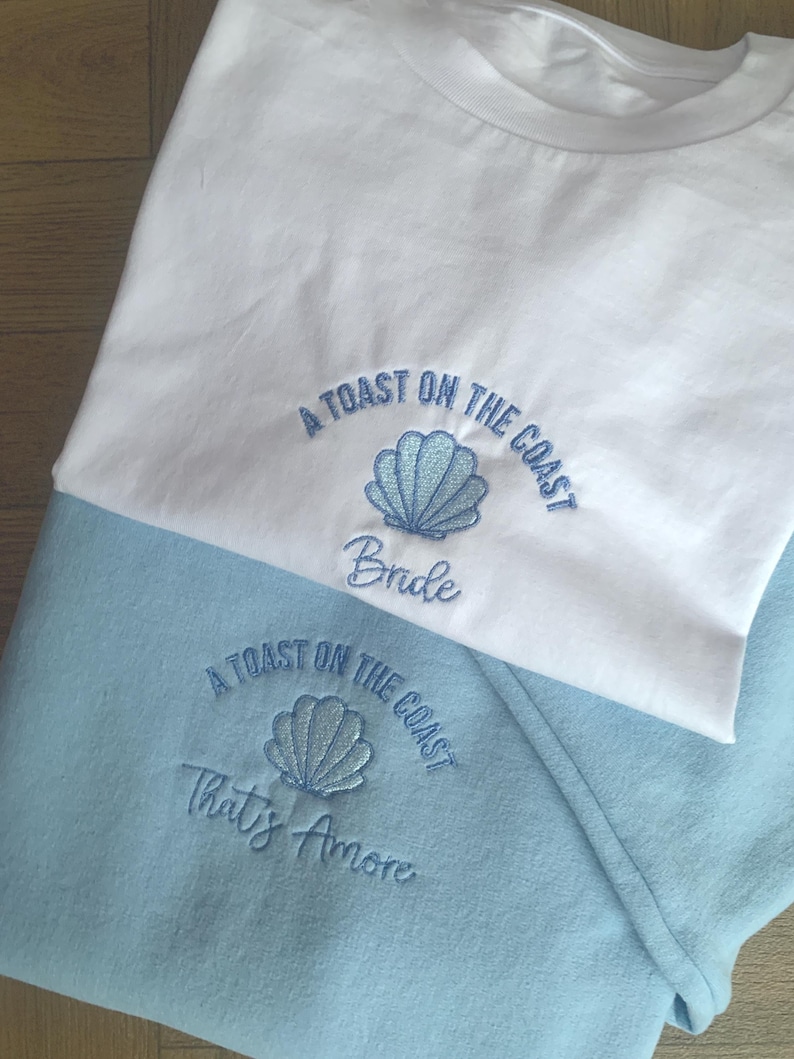 Puede incluir: Camiseta blanca y sudadera azul claro con dise&ntilde;os de conchas bordadas y las frases "A Toast on the Coast" y "Bride" en la camiseta y "That's Amore" en la sudadera. El texto y la concha son de color azul claro.