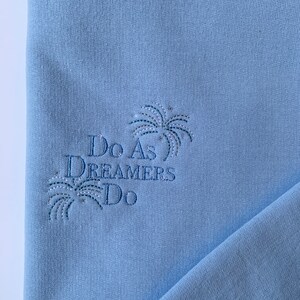Op de afbeelding: Lichtblauwe trui met de tekst "Do As Dreamers Do" geborduurd in donkerblauwe draad. De tekst wordt omringd door een gestileerd vuurwerkontwerp.